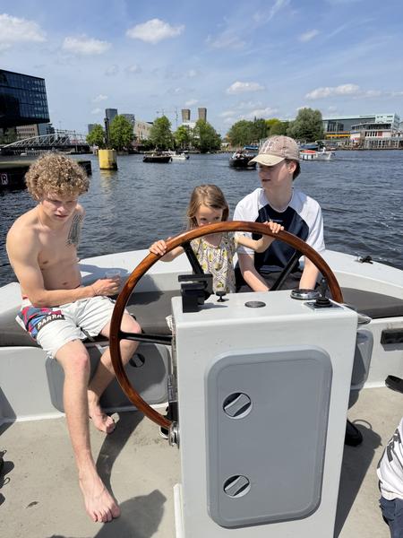 Three individuals are enjoying a sunny day on a boat, with a young girl at the helm, navigating the waters of Bruno van Moerkerkenpassage, Rapenburg, Amsterdam